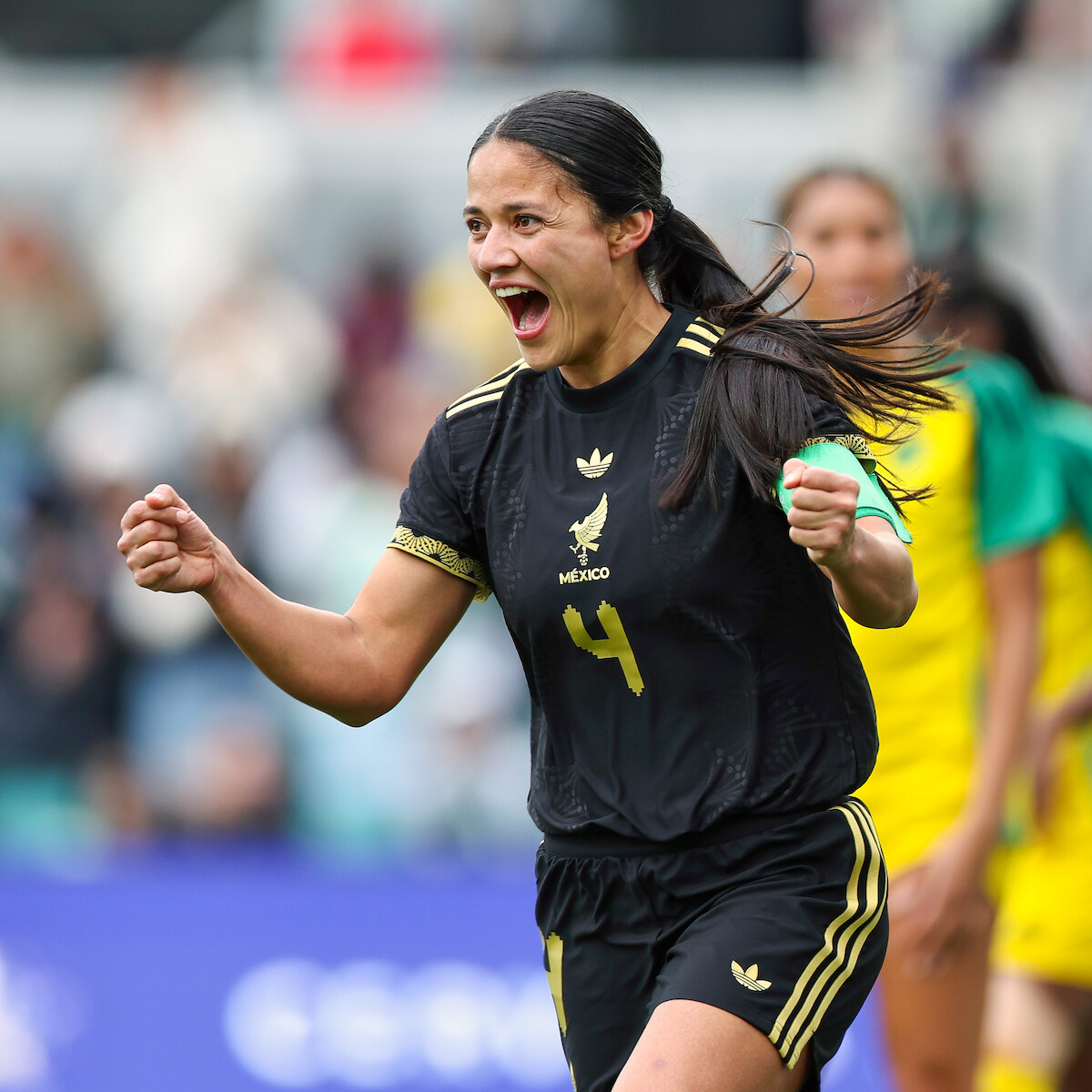Female soccer player in black Mexico uniform celebrating energetically on the field during a match, with blurred teammates and spectators in the background. Outdoor stadium environment, action moment.