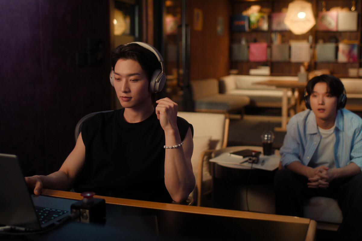 Two people in a cozy, modern room; one in headphones works on a laptop at a desk, the other sits on a sofa with drinks and a notebook on the table, warm lighting and shelves in the background.