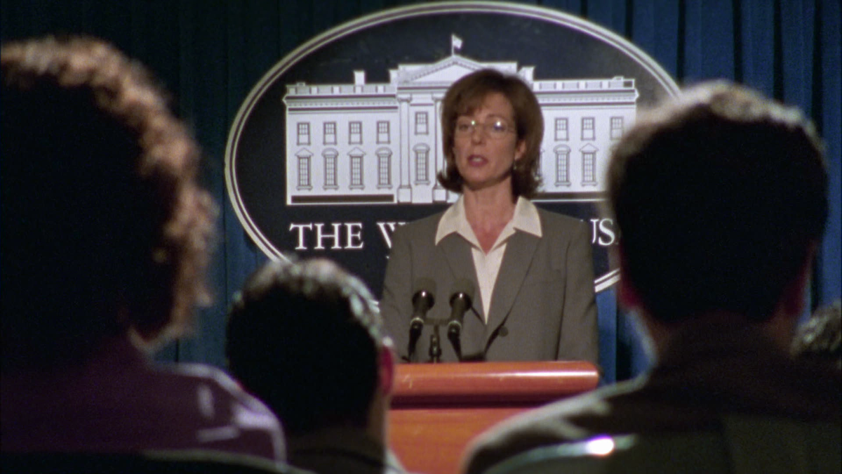 A woman in a suit speaks at a podium with microphones in front of an audience at a White House press briefing, with the White House emblem and curtains in the background.