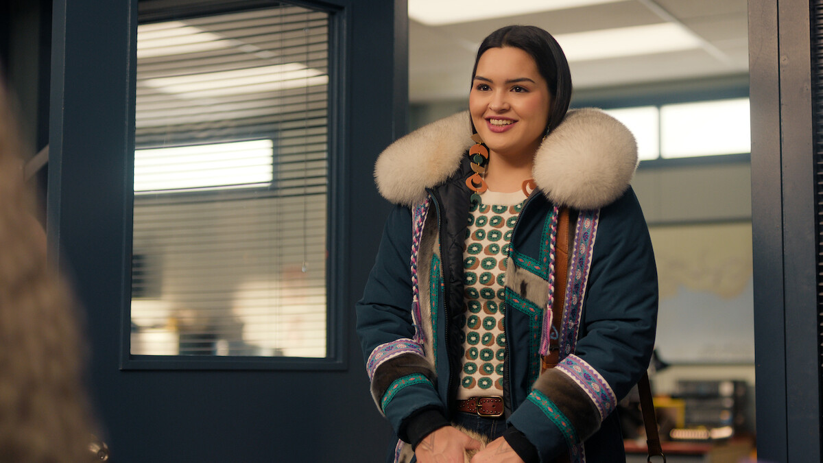 A smiling woman wearing a fur-trimmed parka and patterned shirt stands in an office doorway, with modern office furniture and windows visible in the background.