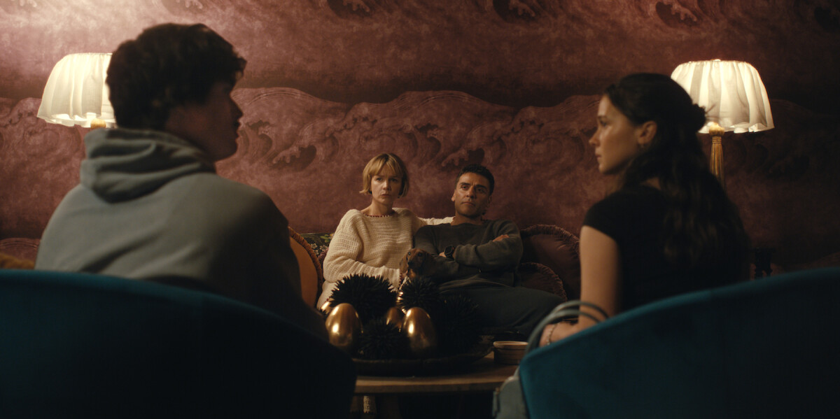 Four people sit in a warmly lit living room with two lamps, plush chairs, and decorative eggs on the table, engaged in an intense conversation against a textured mauve wall.