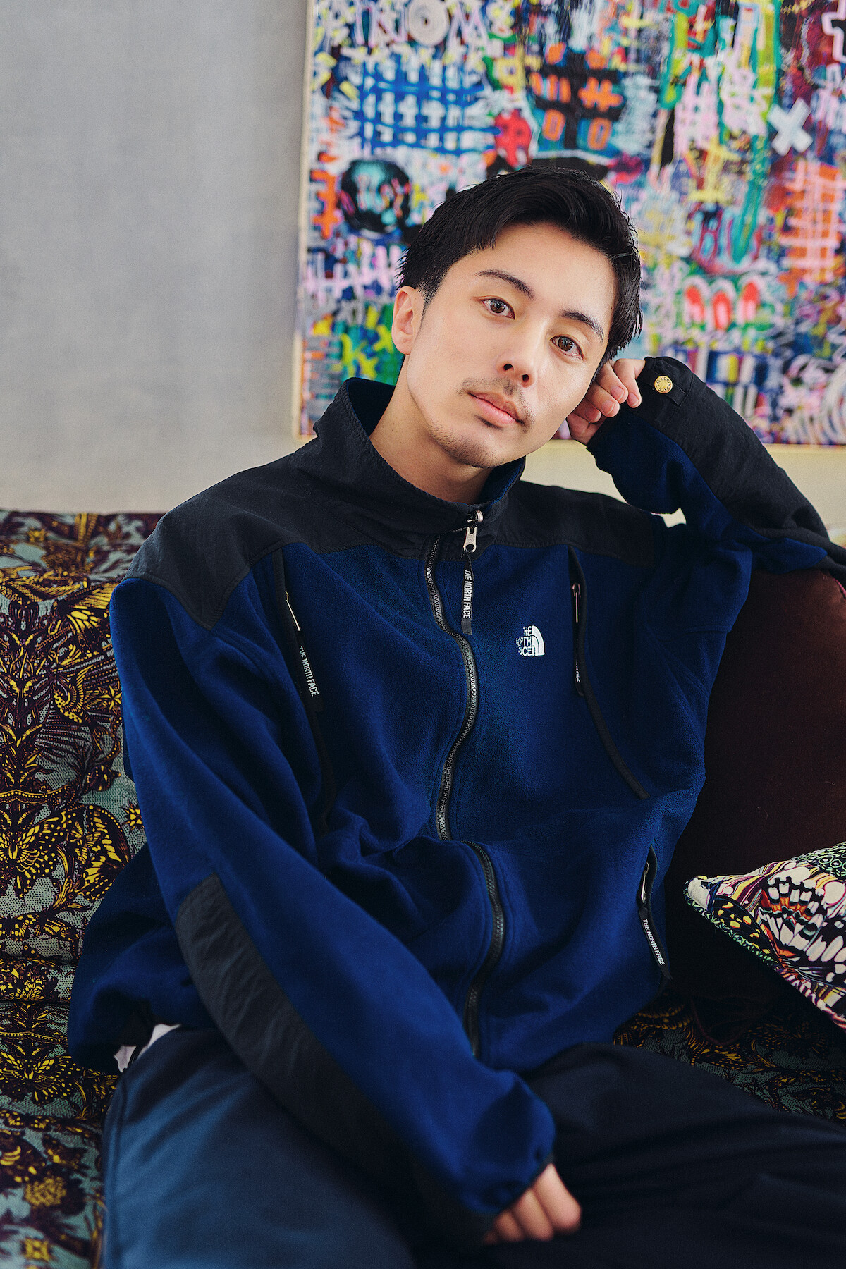 A man in a blue jacket sits on a patterned couch in a cozy room with colorful abstract art on the wall, appearing relaxed and thoughtful.