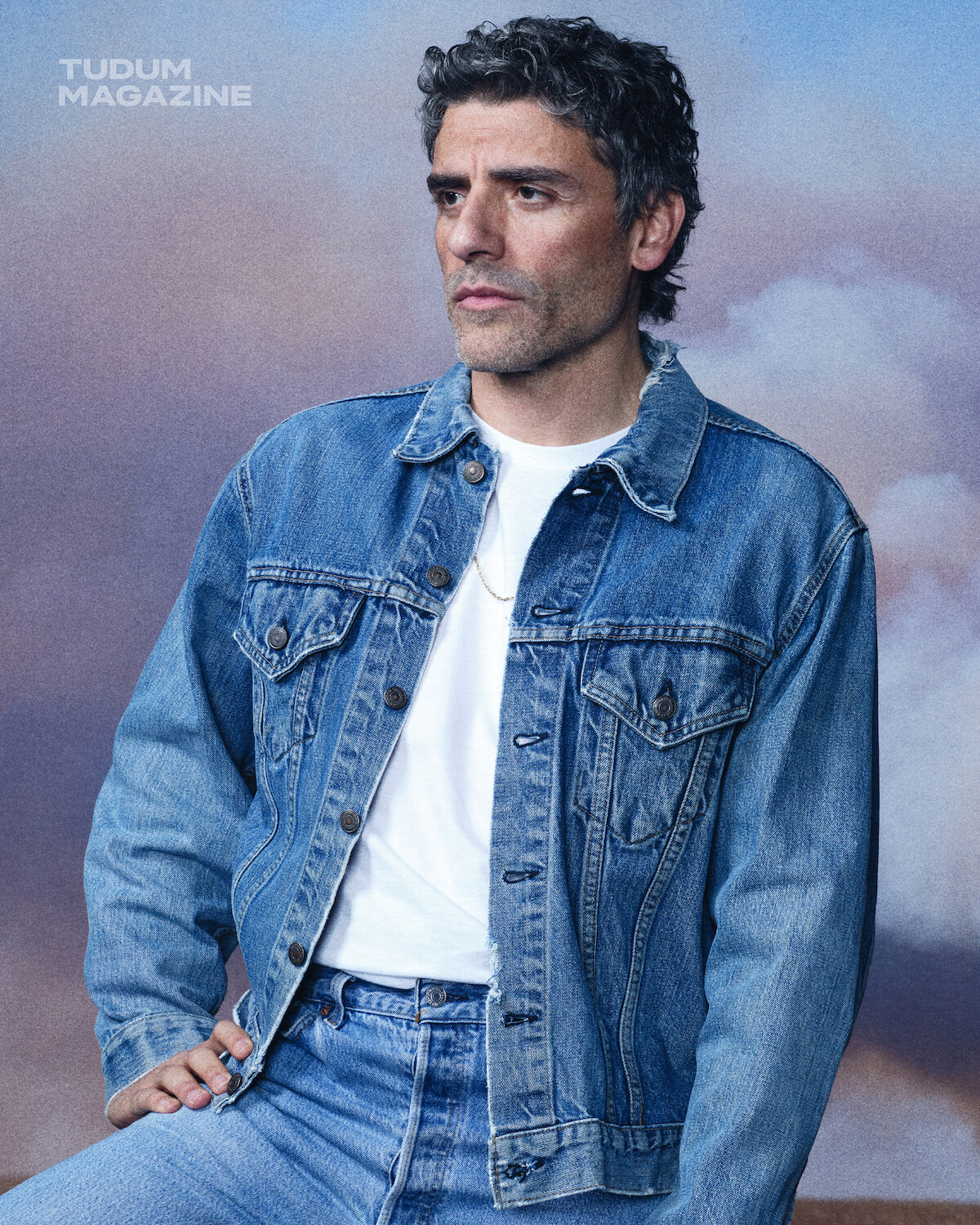 Man wearing blue denim jacket and jeans with a white t-shirt, sitting and posing in front of a cloudy sky backdrop. Studio portrait for Tudum Magazine, looks thoughtful and relaxed in a natural setting.