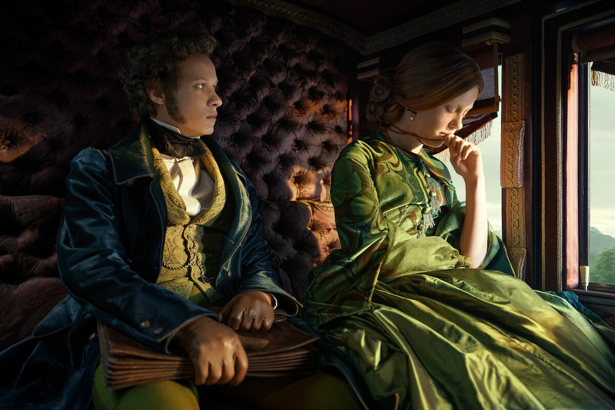 A man and woman in elegant historical clothing sit in a luxurious, tufted train carriage. The woman looks pensively out the window, creating a contemplative and somber mood.