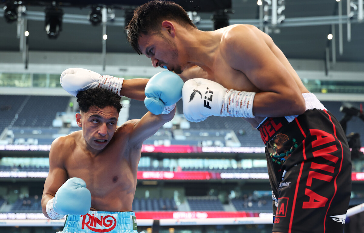 Two male boxers in a match, one in blue trunks and the other in black and red, fighting in a modern indoor boxing arena with spectators and bright lighting in the background.