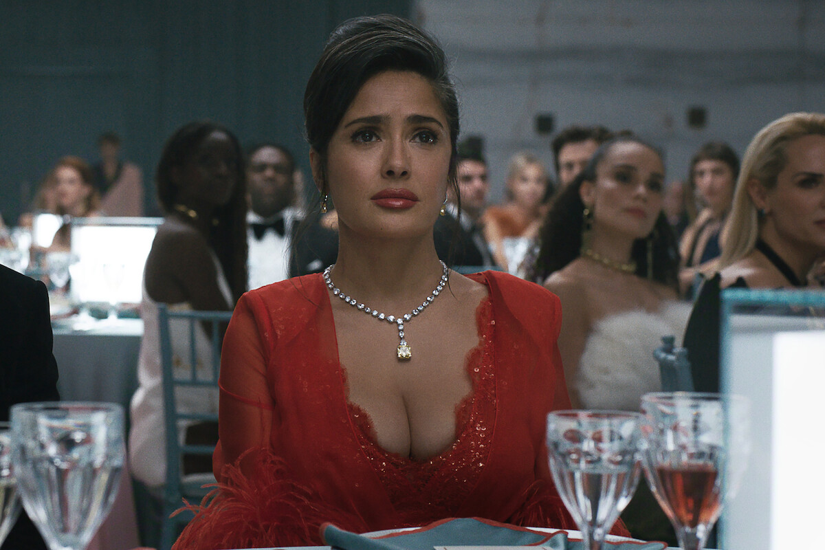 A woman in a red dress with a diamond necklace sits at a formal dinner event, surrounded by elegantly dressed guests at round tables with glassware and plates, in a large, upscale banquet hall.