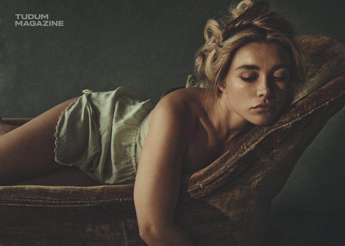 Woman with styled hair wearing silky sleepwear lounges on a vintage chaise in a dim, moody room; soft lighting and muted tones create an intimate, relaxed atmosphere. Tudum Magazine logo appears in the upper corner.