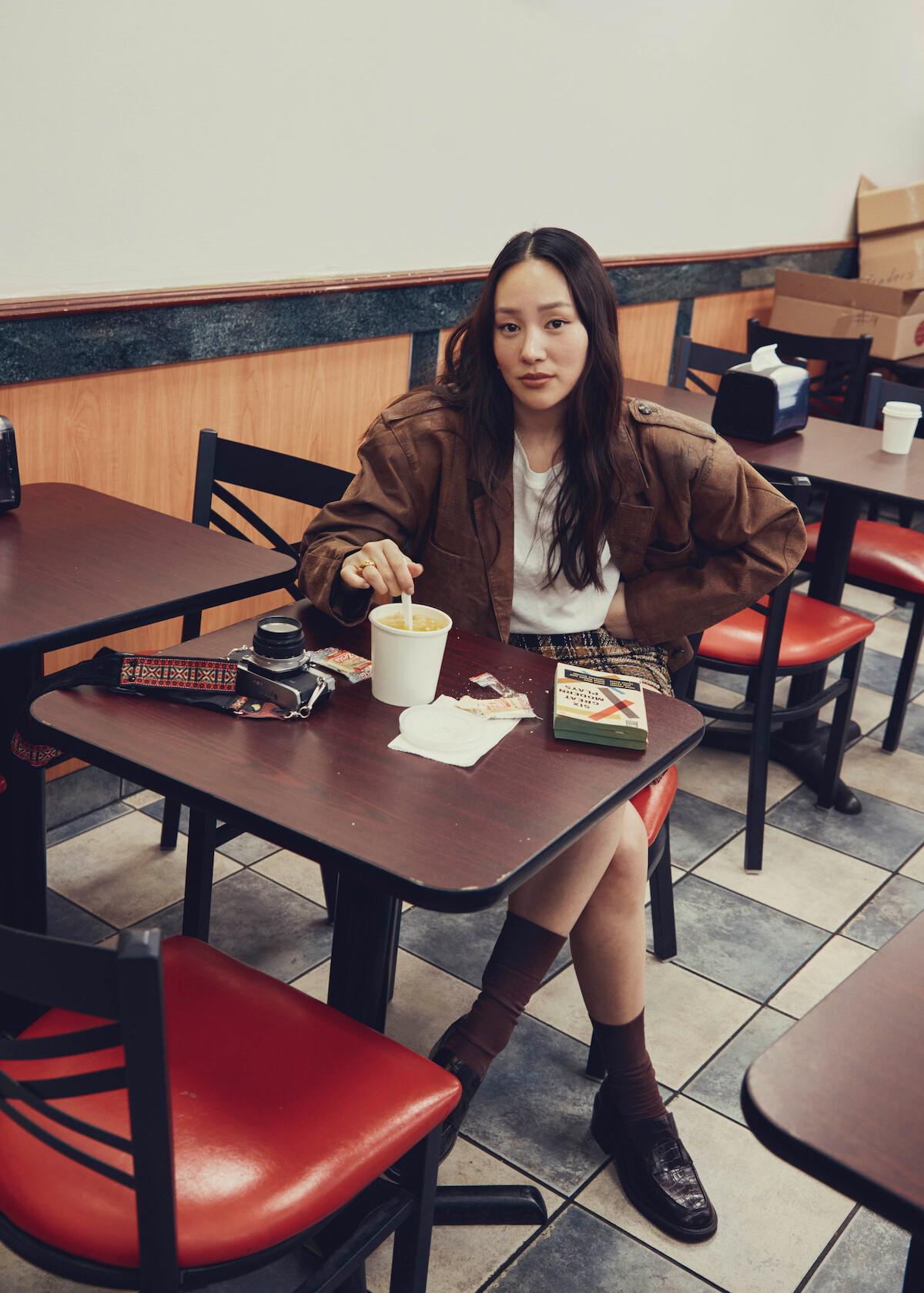 Uma mulher está sentada à mesa em um restaurante simples, comendo macarrão instantâneo no copo. Livros, embalagens de comida, cadeiras vermelhas e um piso de ladrilhos compõem o cenário.
