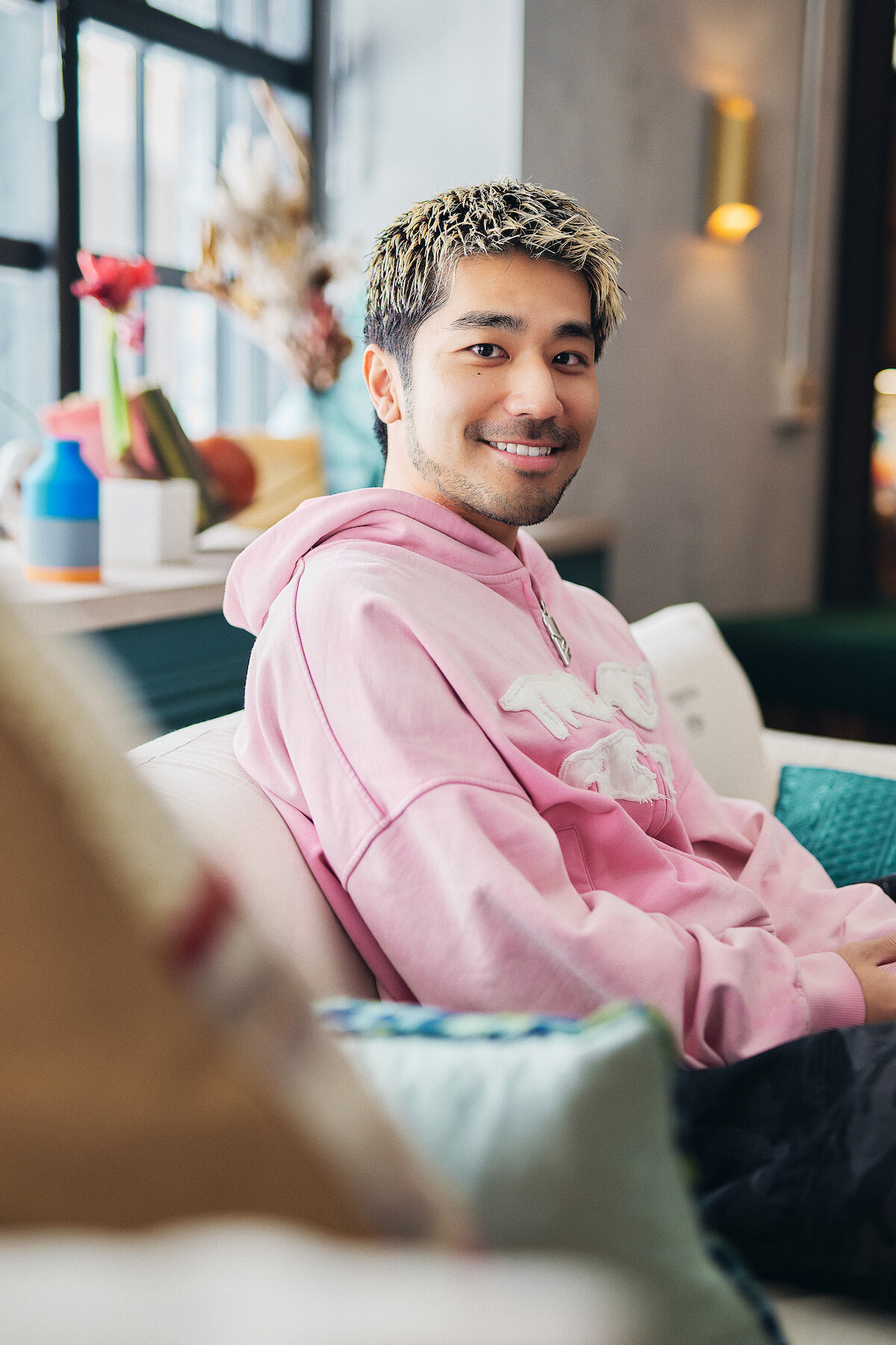 A young man with blond hair in a pink hoodie sits on a couch in a modern, well-lit room with large windows, colorful decor, and a relaxed, friendly atmosphere.
