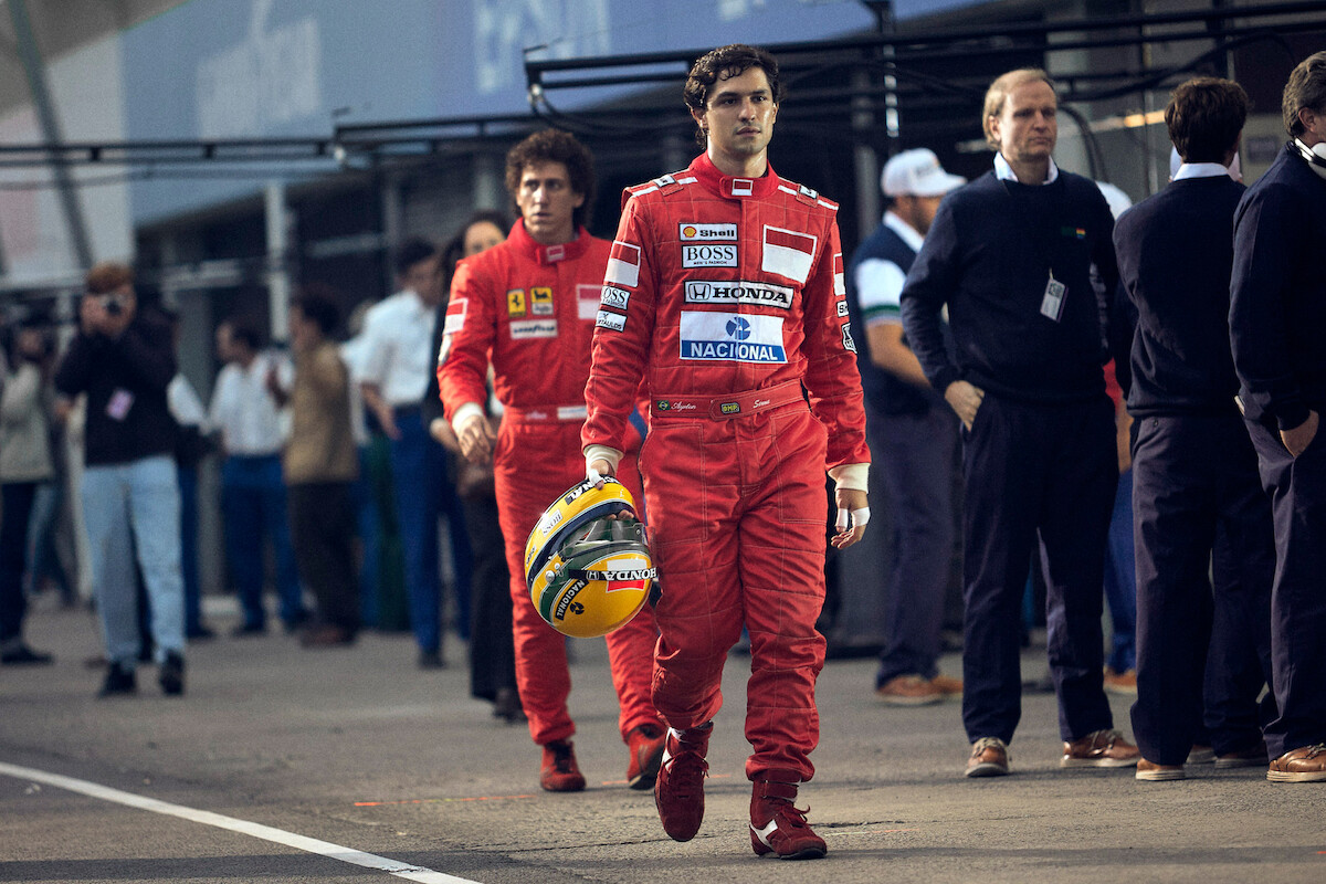 Two race car drivers in red suits walking in a pit lane, one carrying a yellow helmet, with team members and onlookers in the background, capturing a moment of focus before or after a race.