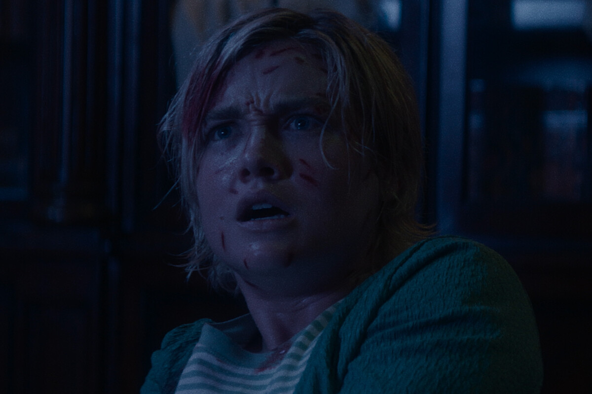 A frightened person with short blond hair and cuts on their face sits indoors in a dimly lit room, wearing a green sweater and striped shirt, looking up with a shocked expression.