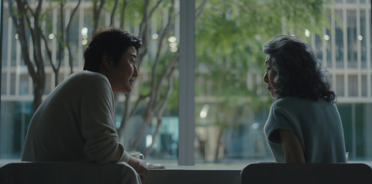 Two people are sitting across from each other at a table in a modern cafe, conversing near a large window with trees and an office building visible outside.