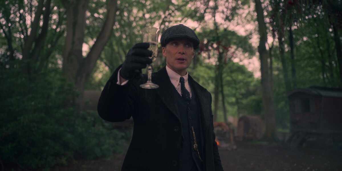 A man in 1920's attire and cap raises a glass of champagne in the woods.