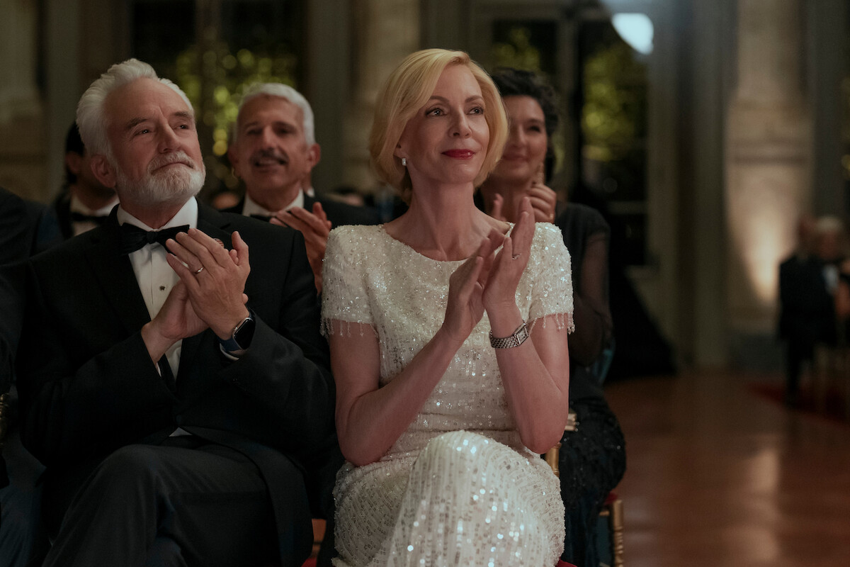 Elegant people in formal attire sit and applaud at an indoor event, possibly a gala or ceremony, with warm lighting and ornate decor in the background.