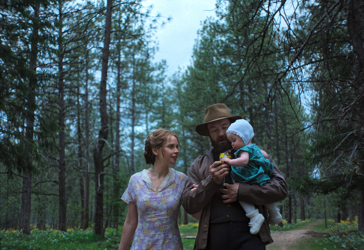 A man and woman walk through a forest, the man gently holding a baby dressed in blue, as wildflowers bloom around them; the mood is serene and intimate, suggesting a family enjoying time together in nature.