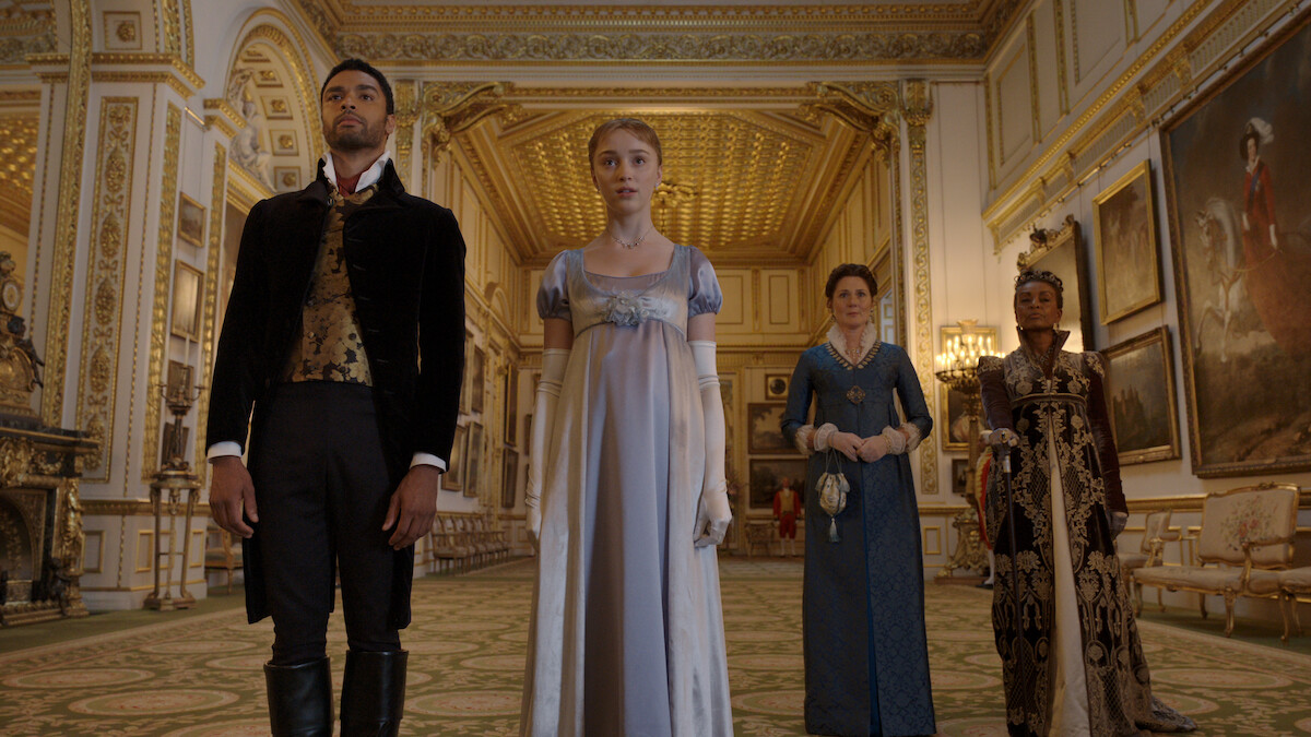 Four people in Regency-era attire stand in an ornate, gold-trimmed ballroom with chandeliers, patterned carpet, and large classical paintings on the walls. The setting suggests a formal or historic event.