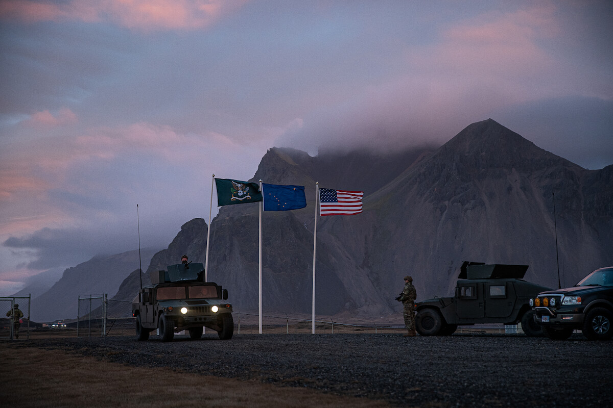 Military vehicles and personnel near three flags at dusk, set against dramatic mountains and a cloudy sky, creating a calm yet vigilant atmosphere in a rugged outdoor environment.