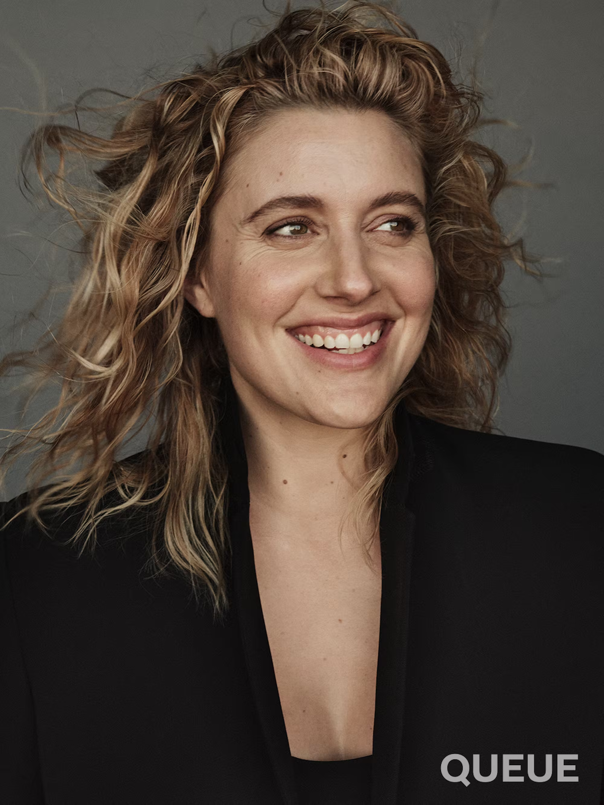 Greta Gerwig smiles against a gray backdrop while wearing a black jacket.