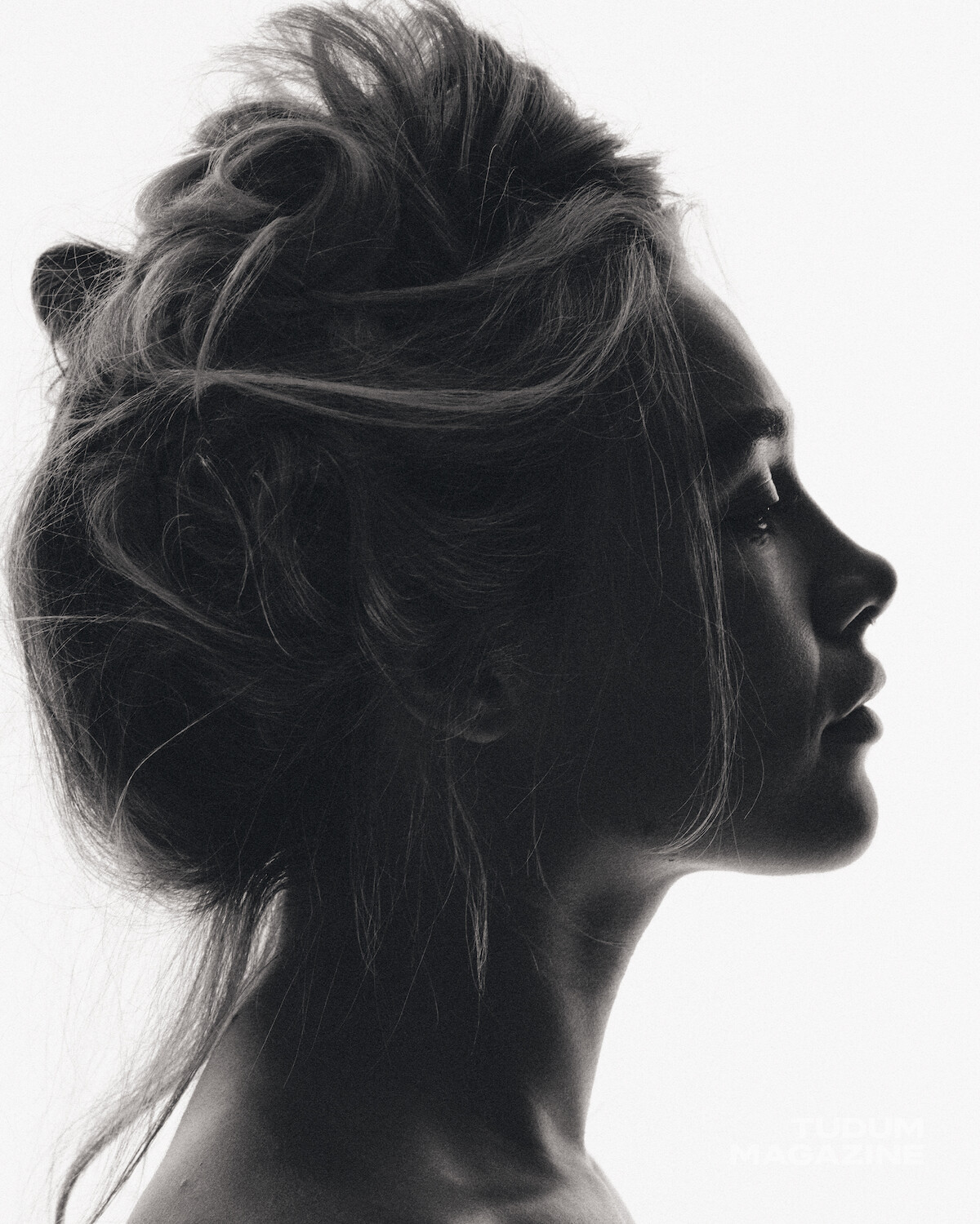 Black and white portrait of a woman in profile with messy hair, styled up. She is against a plain white background, creating a high-contrast, artistic and softly lit setting.