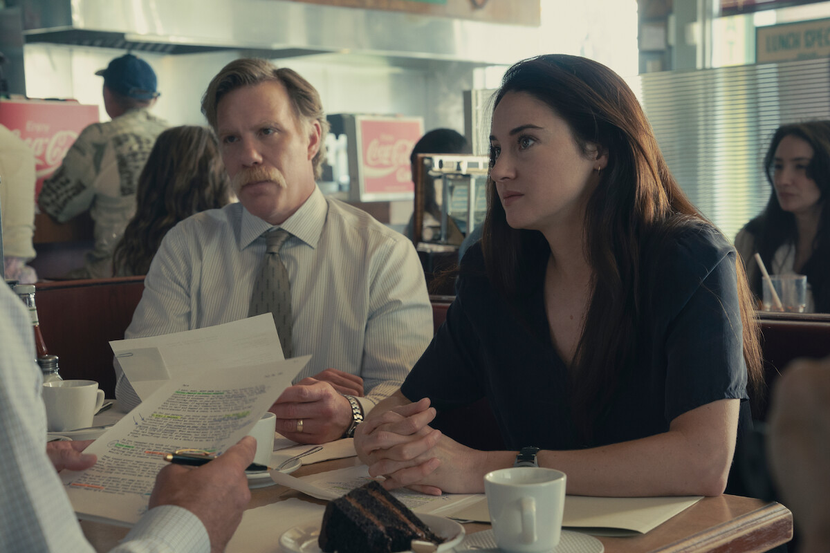 Two people sit at a diner table with coffee and cake, attentively listening to a third person who is holding documents. The setting is casual with retro decor, other patrons are visible in the background.