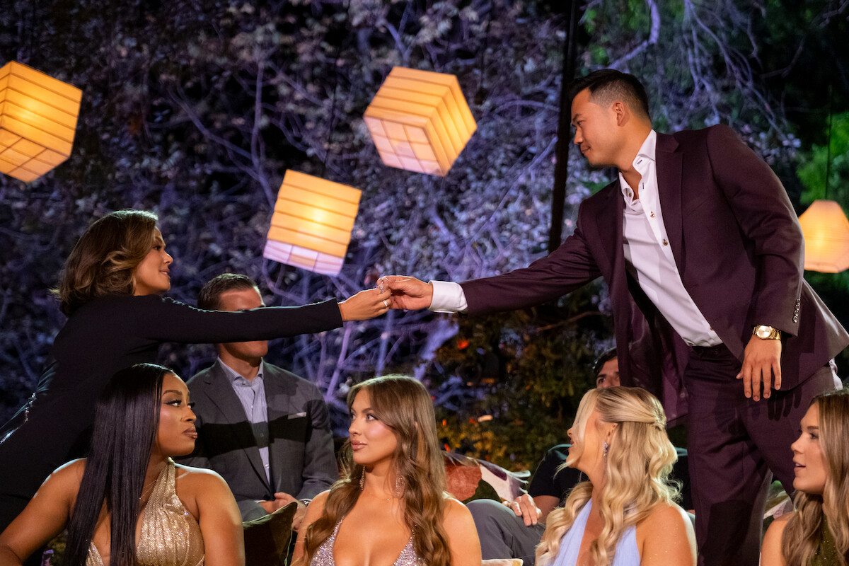 Man in suit reaches to shake hands with seated woman at a formal outdoor gathering, surrounded by other well-dressed people and glowing lanterns, creating a warm, festive, and engaging atmosphere.