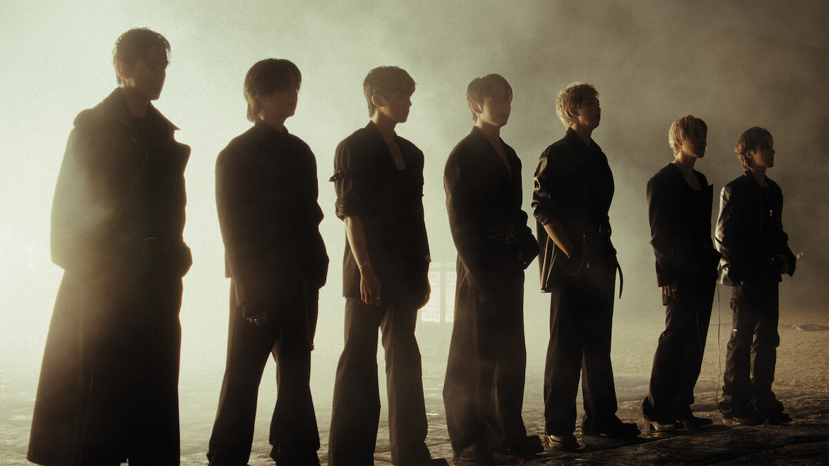 Seven people standing in a row, silhouetted against a bright misty background, wearing dark clothing in a dramatic, moody atmosphere.
