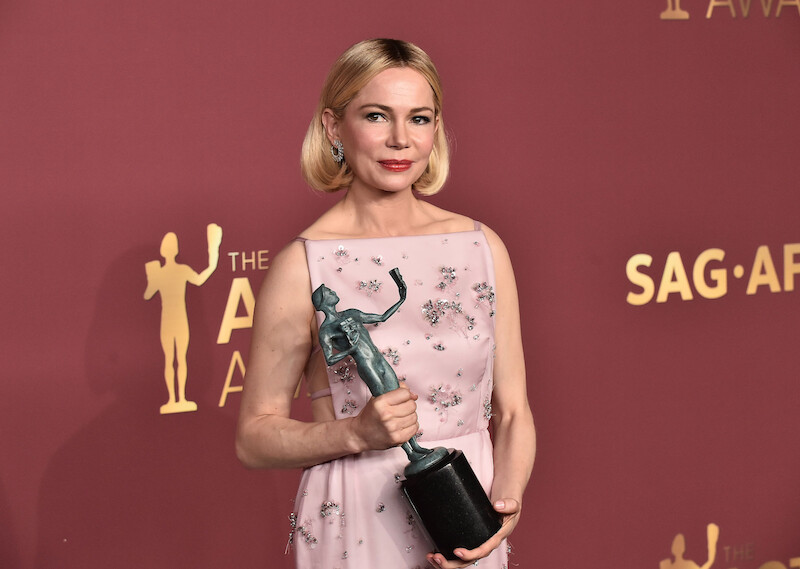 Michelle Williams in pink floral dress holding a trophy at an awards event, standing in front of a red backdrop with gold SAG Awards logos.