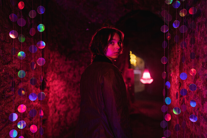 Person standing in a dimly lit tunnel with exposed stone walls, illuminated by vibrant pink, purple, and blue lights and surrounded by hanging colorful circles, creating a mysterious and moody atmosphere.