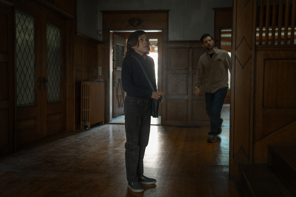 A woman stands in a spacious, dimly lit room with wood paneling, while a man enters from a doorway. The setting appears to be an old house or building with vintage architecture and wooden floors.
