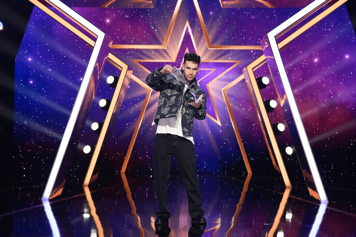 TJ Salta in a gold and black jacket stands center stage with arms open, in front of a large glowing star backdrop and purple, starry lights, likely on a talent or game show set.