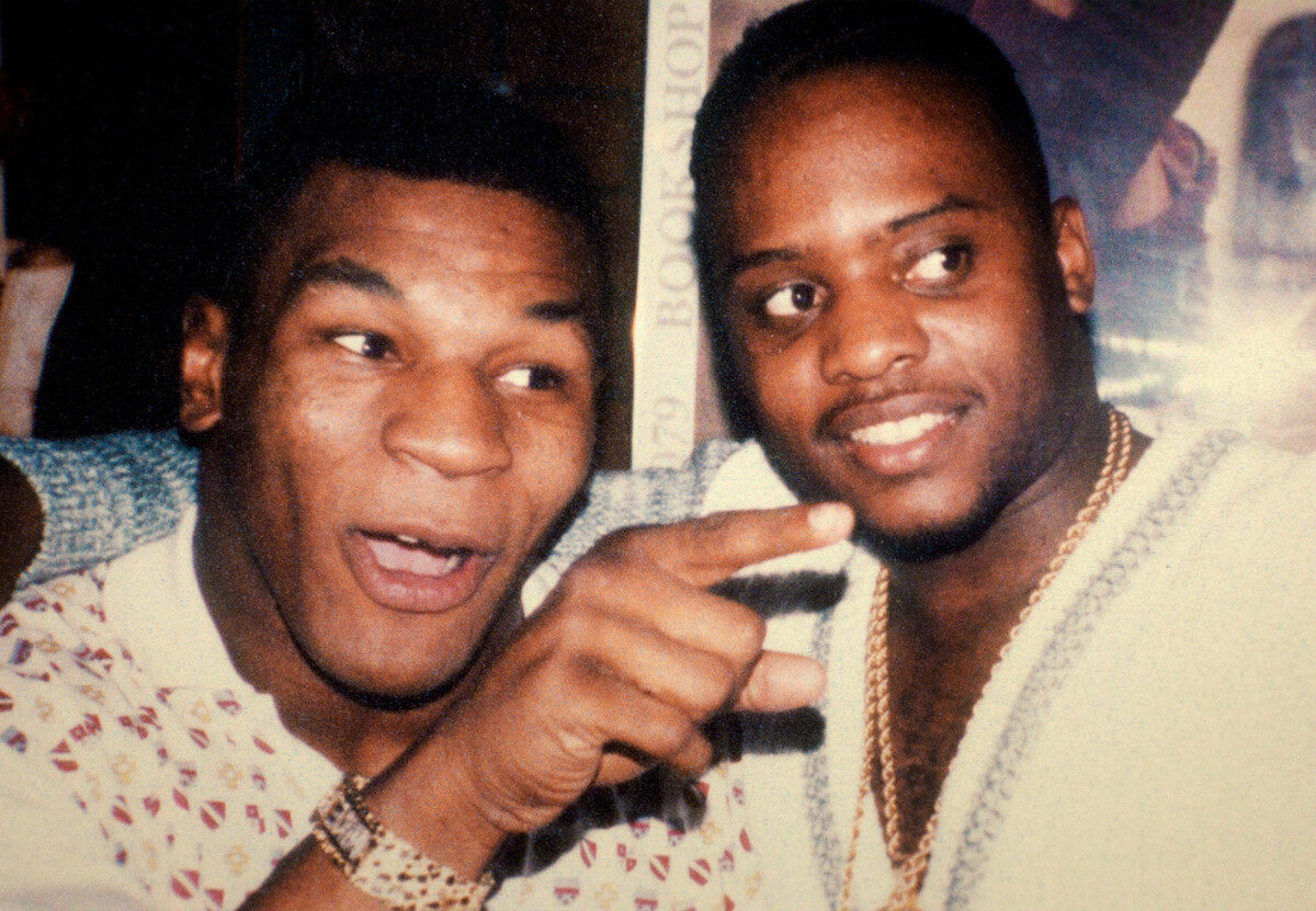 Uma foto antiga mostra Mike Tyson jovem, apontando seu dedo para algo fora da imagem e sorrindo