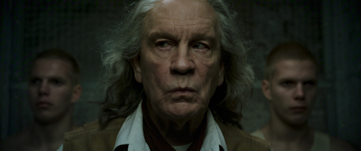 Older man with long gray hair stands in the center, looking serious, flanked by two young men with shaved heads. The setting is dimly lit, with a gritty, industrial or prison-like background.