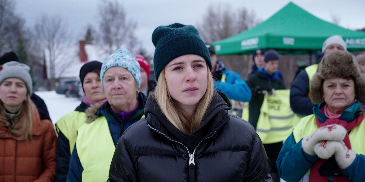 Carla Sehn wears a black jacket and black beanie while standing outside in front of a group of people in ‘The Åre Murders.’
