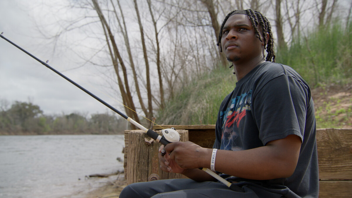 Young man fishing by a riverbank, sitting on a wooden bench surrounded by trees and greenery on a cloudy day, holding a fishing rod and looking thoughtful.