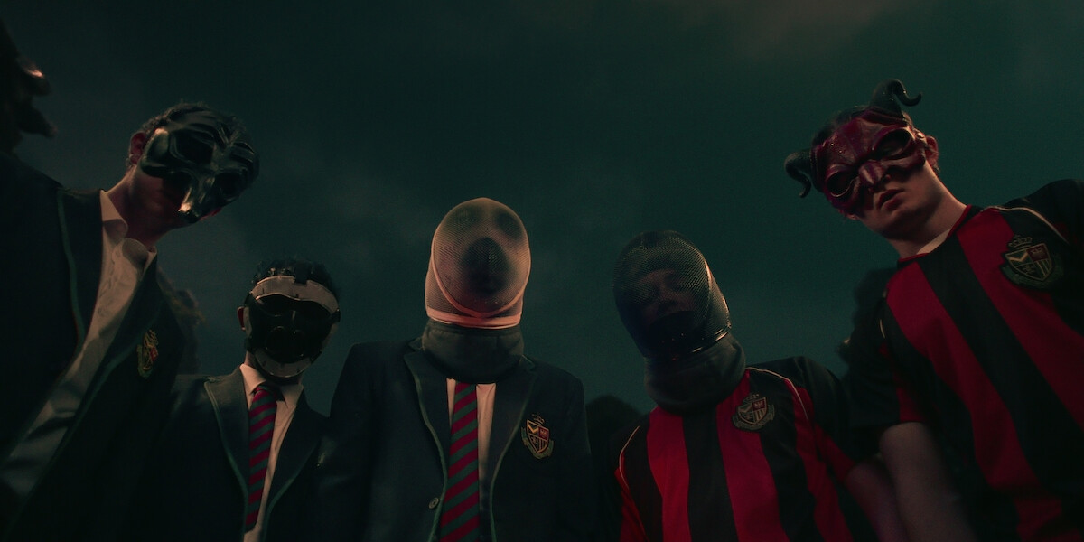 A group of five men wearing masquerade and fencing masks stand together looking down in the series ‘Fool Me Once.’