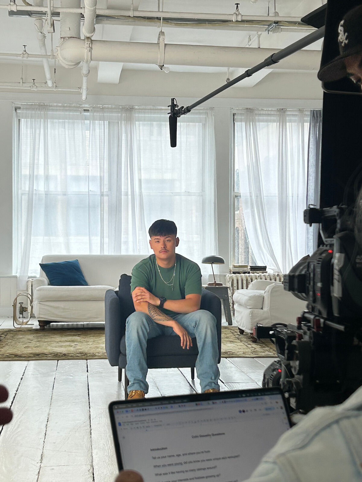 Young man sits on a chair in a bright, modern living room with white sofas and large windows. Filming equipment, camera, microphone, and someone holding a laptop are visible, indicating a video interview or production setting.