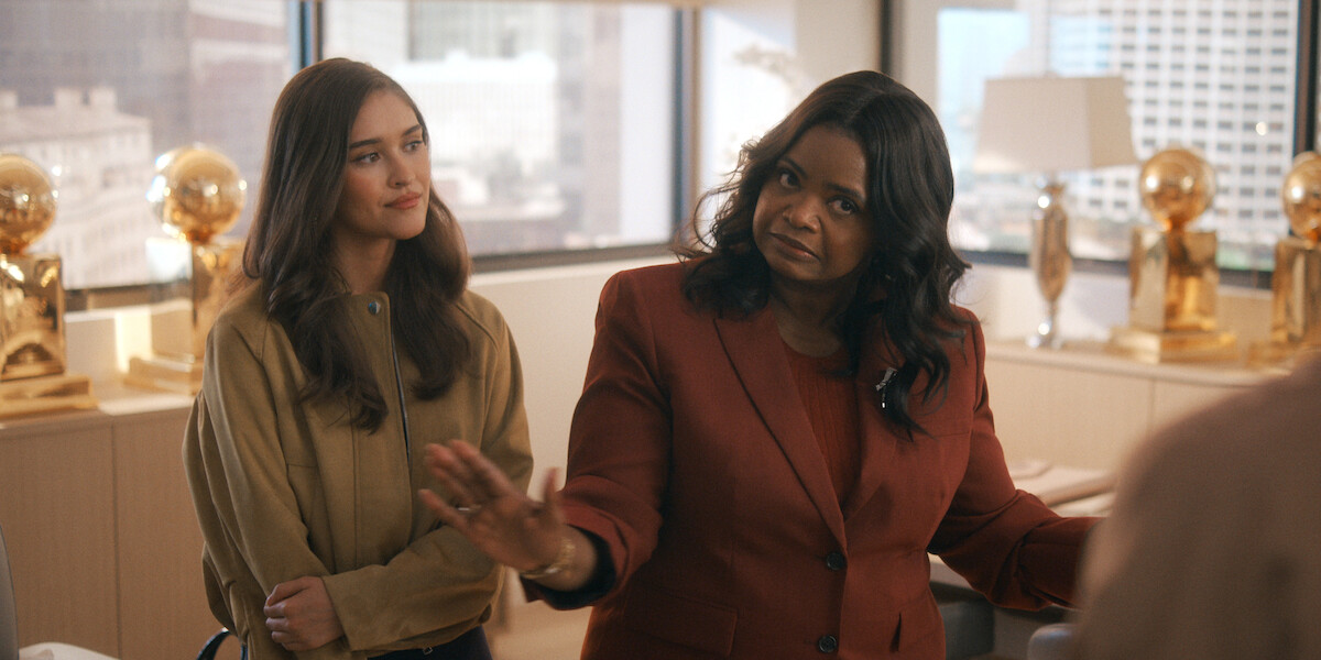 Two women standing in a modern office with large windows and several golden basketball trophies on display in the background, one gesturing with her hands, the other observing calmly.