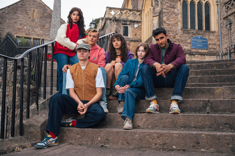 Six young people sit and pose on historic stone steps outside an old church building, dressed in colorful casual outfits, creating a moody and thoughtful atmosphere in a picturesque outdoor setting.