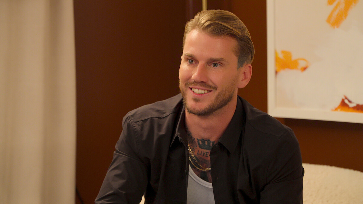Smiling man with blond hair and visible chest tattoos wearing a black shirt sits indoors on a chair, with warm brown walls, a curtain, and abstract art in the background.