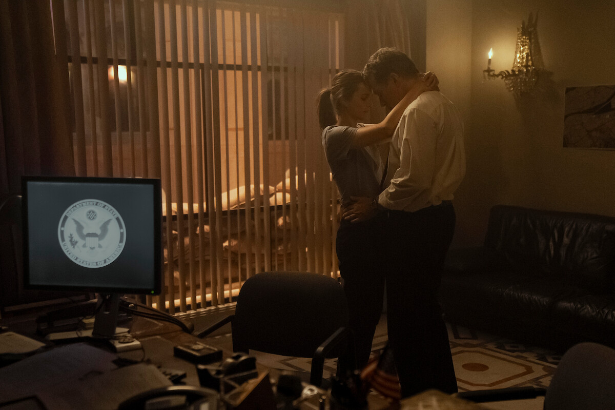 A man and woman embrace in a dimly lit office with a computer screen, desk, and couch. Blinds and warm lighting suggest an intimate, secretive setting.