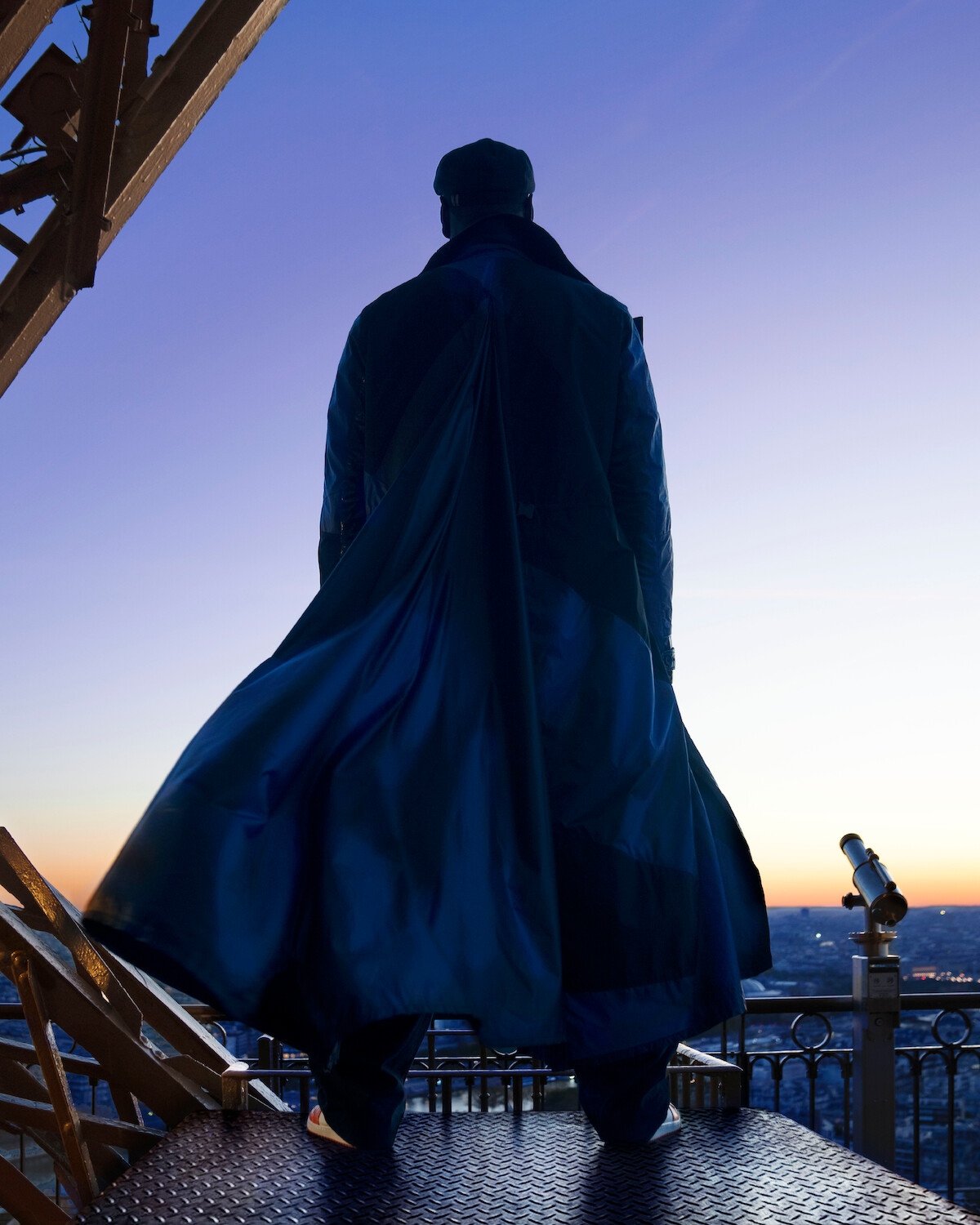 Um homem aparece de costas, vestindo um sobretudo azul-marinho e chapéu, em cima da torre Eiffel