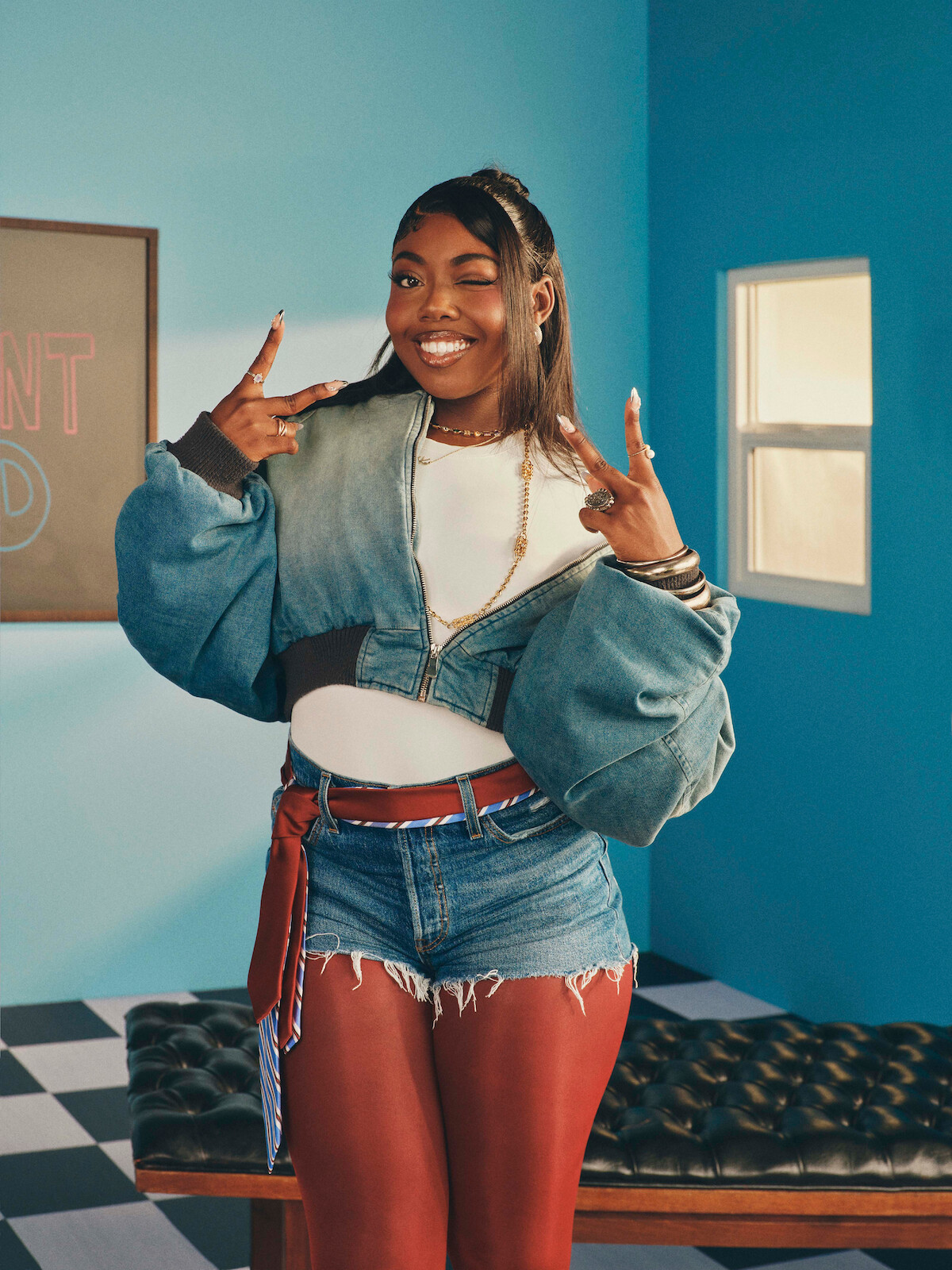 Smiling woman in a denim jacket and shorts poses indoors, making peace signs with both hands. She stands in a bright room with blue walls, a window, checkered floor, chalkboard, and tufted bench.