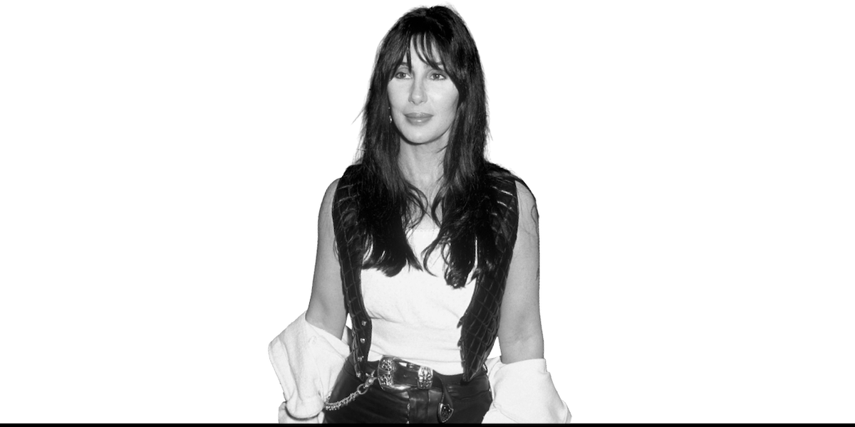 A woman with long dark hair stands facing the camera, wearing a white shirt, dark vest, and belt. The image is in black and white with no visible background.