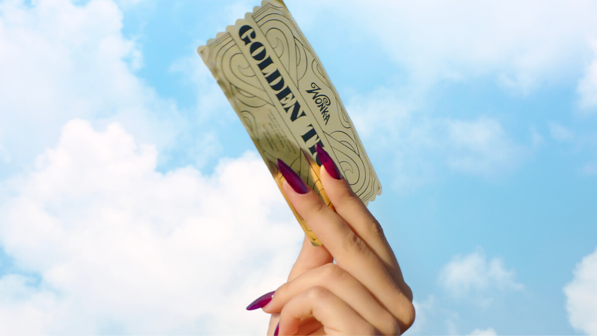A hand with long, painted nails holds up a golden ticket against a blue sky with clouds.