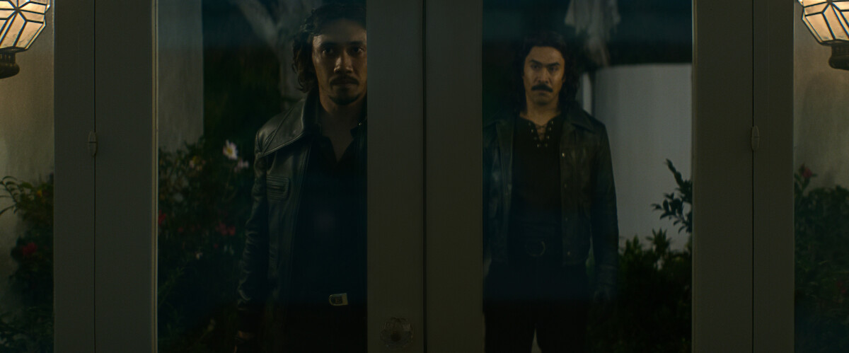 Two men standing outside at night, seen through glass doors with reflections, both wearing dark jackets, with outdoor wall lamps and plants visible, giving a tense or mysterious atmosphere.