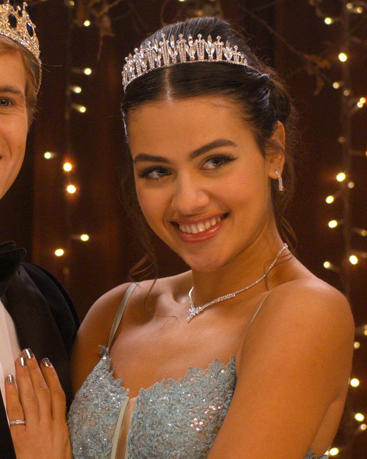 Young woman wearing a sparkling tiara and light blue gown, smiling at a formal event with elegant lighting and festive string lights in the background.