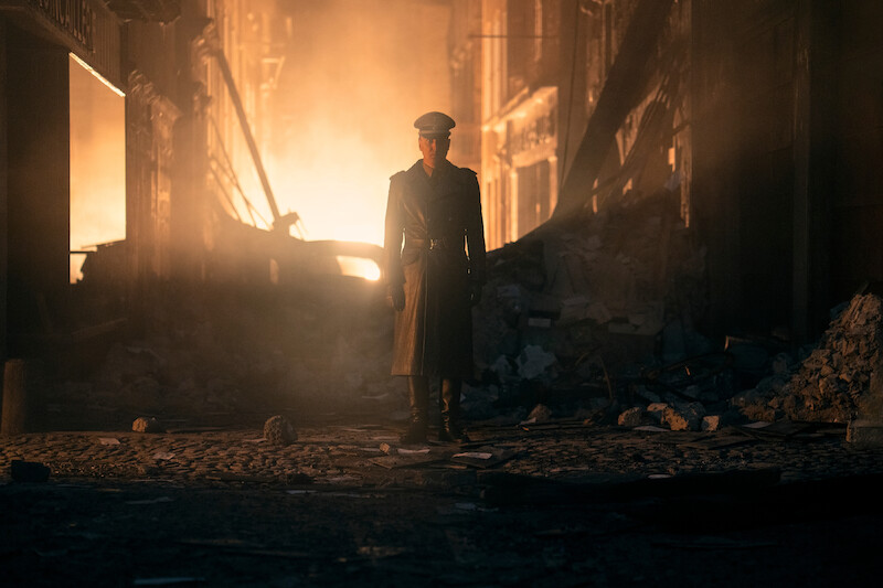 Lars Eidinger as Sergeant Major Reinhold von Rumpel standing in a ruined street. 