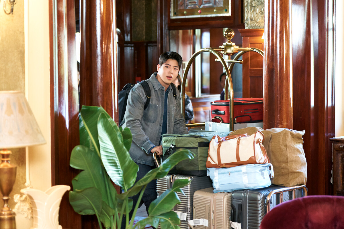 Young man in a hotel lobby with a backpack, standing beside a luggage cart loaded with suitcases and bags amid polished wood columns and lush indoor plants.