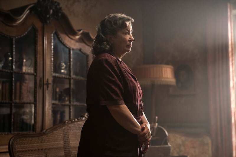Marion Bailey as Madame Manec stands in a sitting room, with a large cabinet and wicker bench behind her. 