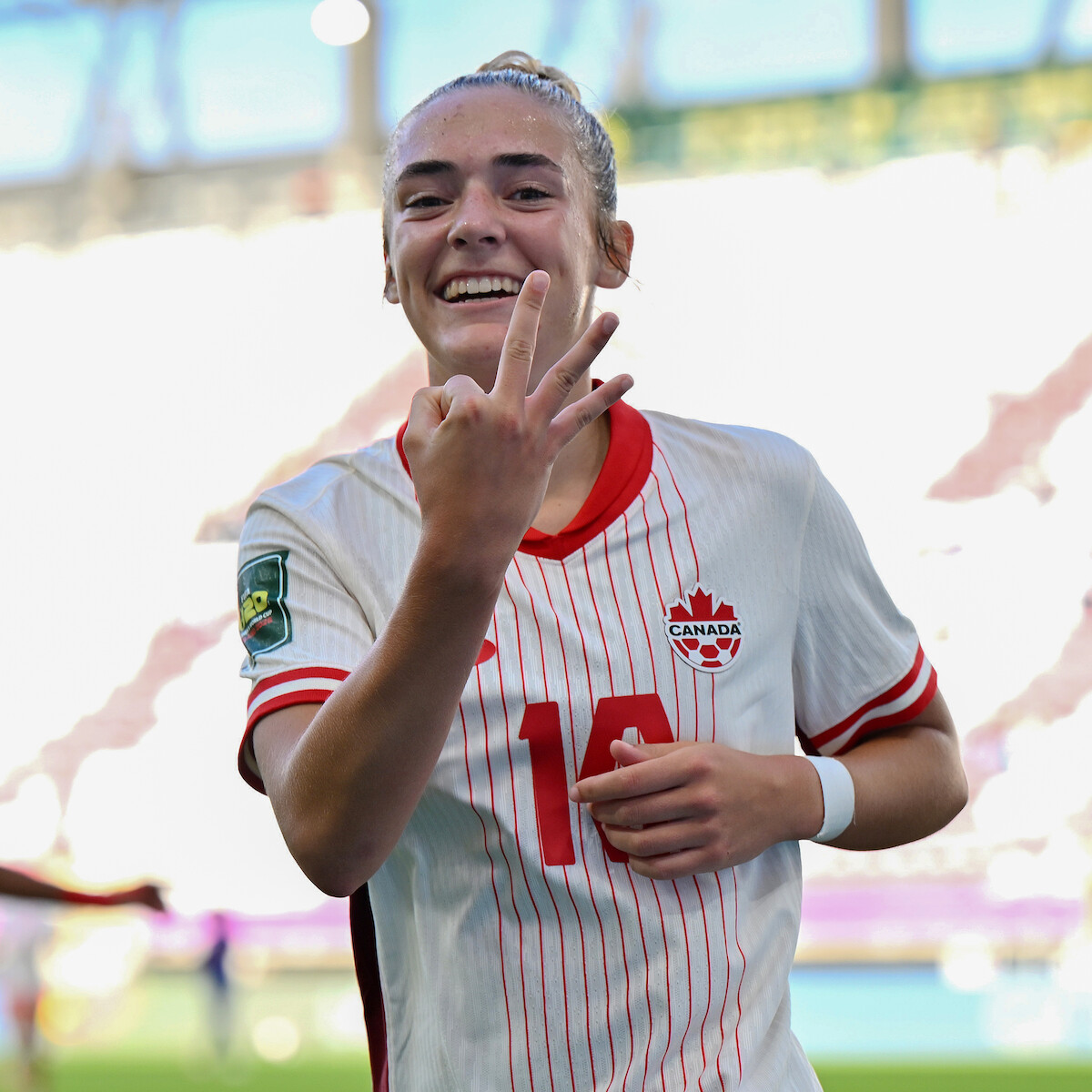 Female soccer player in a white Canada jersey smiling, holding up three fingers, standing on a soccer field in a stadium with bright lighting and blurred audience in the background.